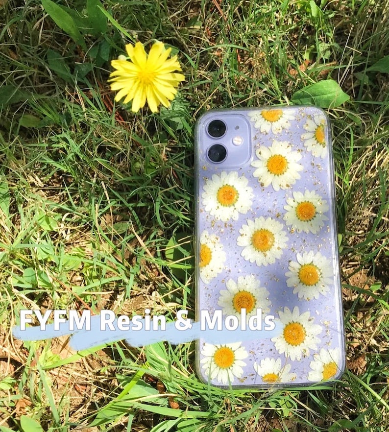 May include: A clear phone case with a purple background and a pattern of white daisies and gold glitter. The case is lying on a bed of green grass.