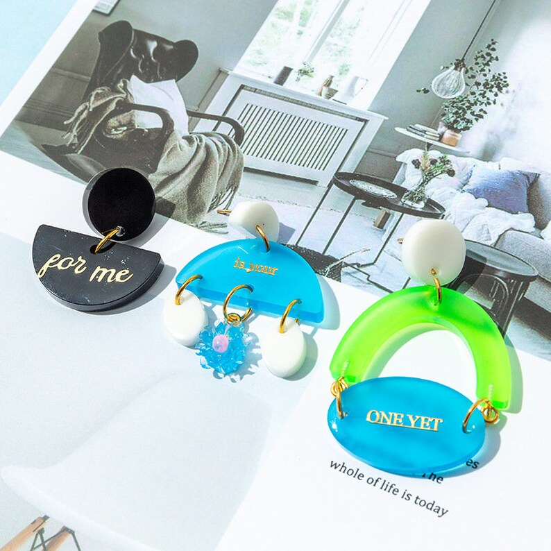 May include: Three pairs of colorful resin earrings with gold accents. The earrings feature a black half-circle with the text "for me", a blue half-circle with the text "is yours", and a green arch with a blue oval that says "one yet whole of life is today".