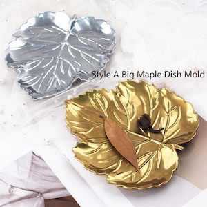 May include: Two decorative leaf-shaped dishes. One is silver, the other is gold. The gold dish holds a dried leaf and a small, dark brown object. The text "Style A Big Maple Dish Mold" is visible.