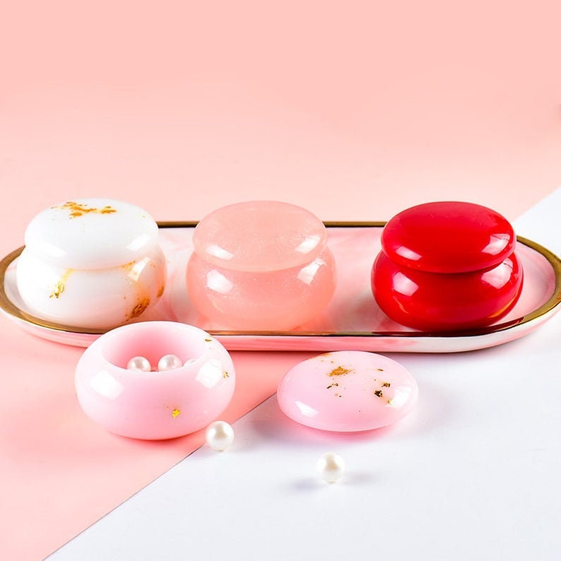 May include: Four small, round, decorative boxes with lids. The boxes are in shades of pink, white, and red. The boxes are on a white surface with a pink background. There are three small white pearls scattered on the surface.