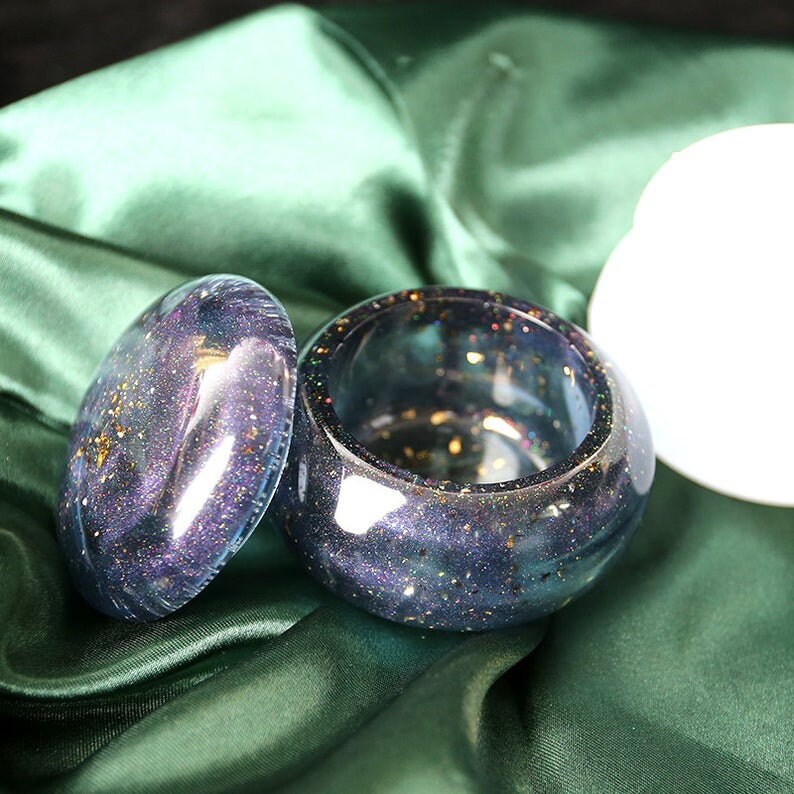 May include: A round, iridescent blue and purple resin trinket box with a lid. The box is decorated with gold glitter and has a speckled appearance. The box is sitting on a green fabric surface.