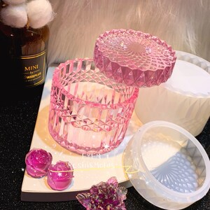 May include: A pink, faceted resin container with a matching lid, alongside a white mold and rose-shaped resin pieces. The container is open, revealing a white interior. The text "FYFM Resins Molds" is visible.