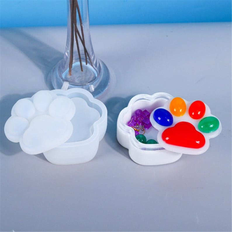 Pu&ograve; includere: Due stampi in resina a forma di impronta di zampa bianca. Uno stampo &egrave; aperto e mostra un design di impronta di zampa multicolore all'interno. L'altro stampo &egrave; chiuso.