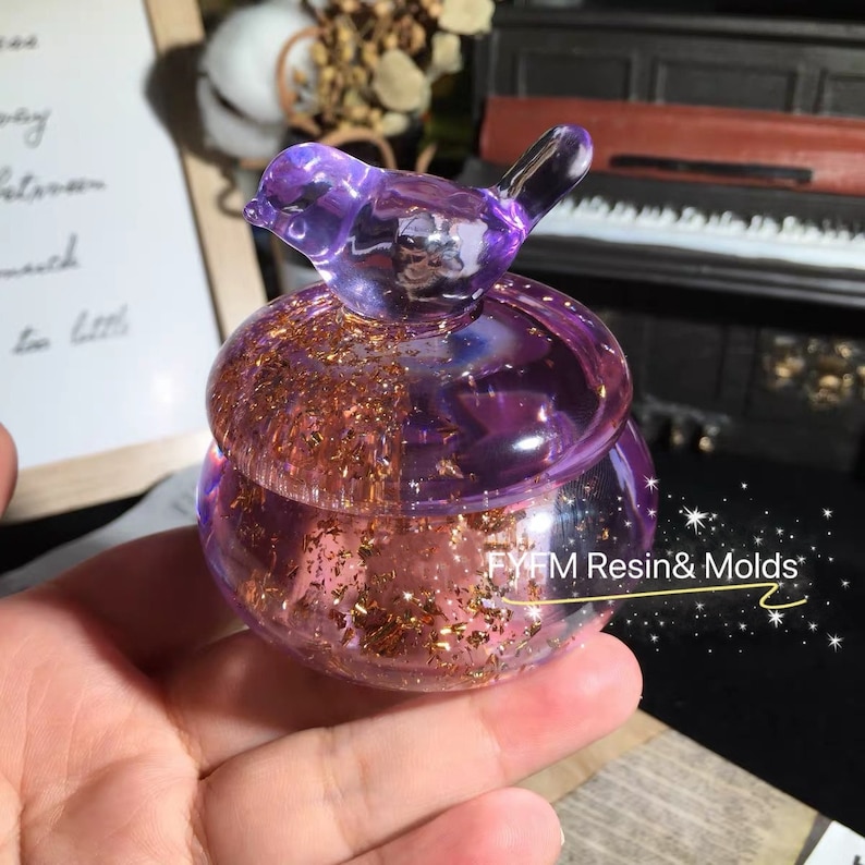 May include: A purple glass jar with a bird-shaped lid. The jar is filled with gold glitter and has a pink resin base. The jar is sitting on a hand.