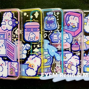 May include: Four clear phone cases with colorful cartoon designs. Each case features a different design with a cute bunny, a cloud, a bottle of soap, and a kitchen appliance. The cases are all made of clear plastic and have a glossy finish.