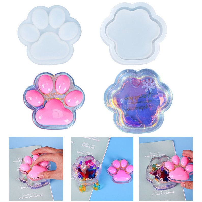 Pu&ograve; includere: Due stampi in silicone trasparente a forma di impronta di zampa, uno riempito con resina rosa e glitter, l'altro con resina blu e glitter. Gli stampi sono su uno sfondo bianco.