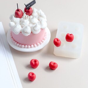 Op de afbeelding: Een roze cake met witte frosting en rode kersen erop. De cake staat op een wit bord met een parelrand. Naast de cake liggen vijf rode kersen in een siliconen mal. De mal heeft negen holtes.