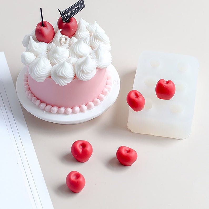 Op de afbeelding: Een roze cake met witte frosting en rode kersen erop. De cake staat op een wit bord met een parelrand. Naast de cake liggen vijf rode kersen in een siliconen mal. De mal heeft negen holtes.