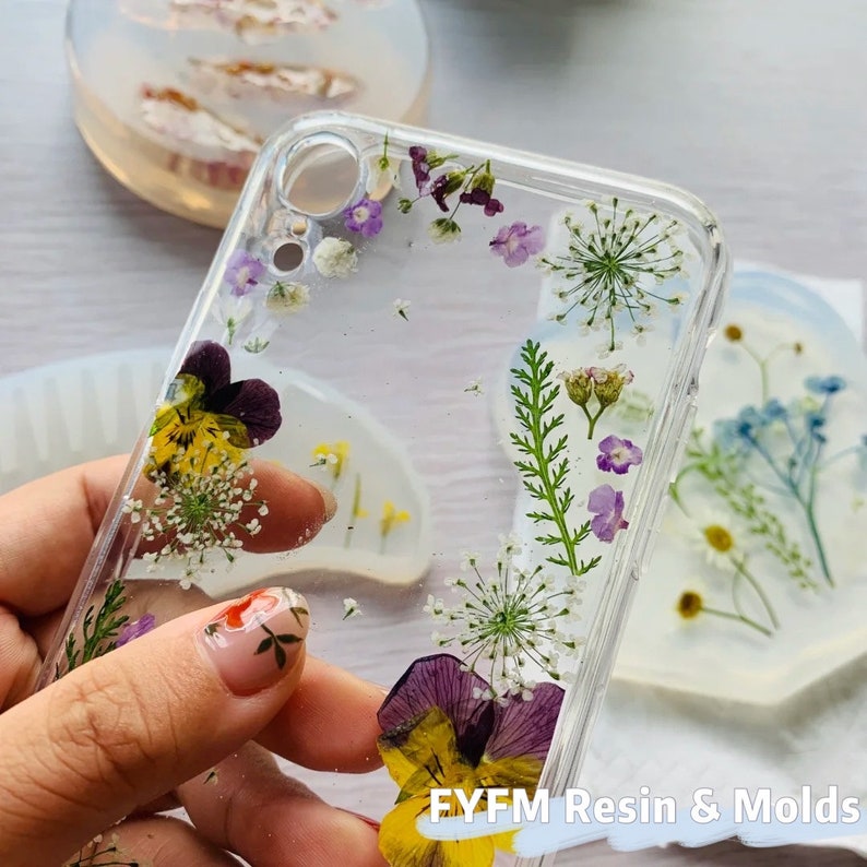 May include: A clear phone case with pressed flowers and greenery inside. The flowers are purple, yellow, and white. The case is made of resin and has a clear, glossy finish. The text "FYFM Resin & Molds" is visible at the bottom of the image.