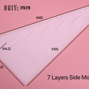 Pode incluir: Um molde de pl&aacute;stico transparente para fazer 7 camadas de bolo. O molde tem a forma de um tri&acirc;ngulo com medidas em mil&iacute;metros. O texto "UNIT: mm" e "7 Layers Side Mold" est&aacute; impresso no molde.