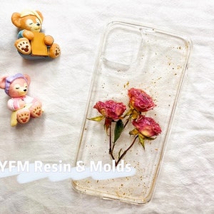 May include: Clear phone case with a gold glitter background and pressed pink flowers. The case is designed for a smartphone with a camera cutout.