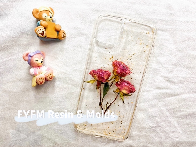 May include: Clear phone case with a gold glitter background and pressed pink flowers. The case is designed for a smartphone with a camera cutout.