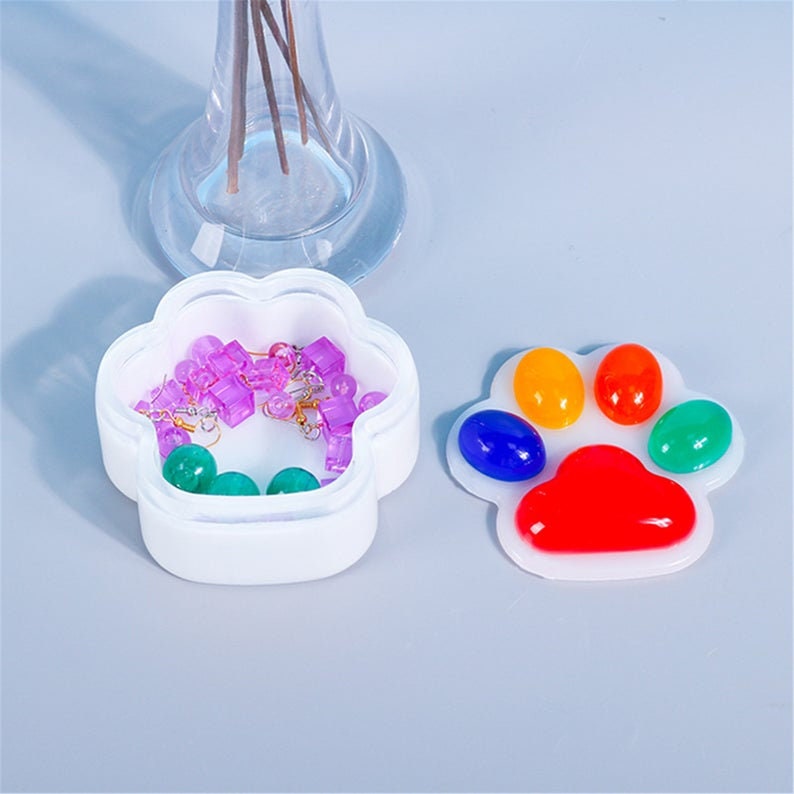Pu&ograve; includere: Due contenitori bianchi a forma di zampa. Un contenitore &egrave; aperto e riempito con perline viola e verdi e una catena dorata. L'altro contenitore &egrave; chiuso e ha un design a forma di impronta di zampa arcobaleno sul coperchio.