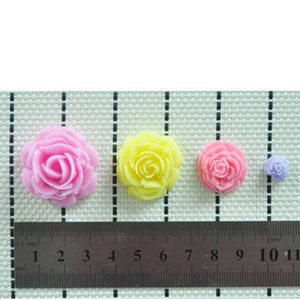 May include: Four rose-shaped decorations in pink, yellow, pink, and purple. A ruler with measurements in centimetres is placed below the flowers, showing their relative sizes. The flowers are arranged on a textured surface.
