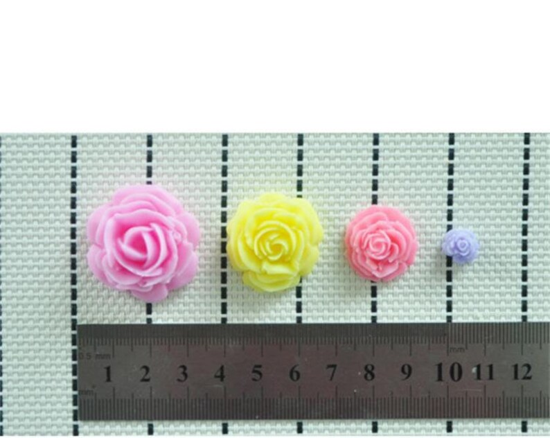 May include: Four rose-shaped decorations in pink, yellow, pink, and purple. A ruler with measurements in centimetres is placed below the flowers, showing their relative sizes. The flowers are arranged on a textured surface.