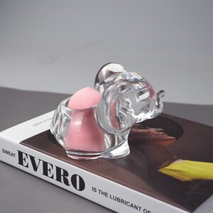 Puede incluir: Una figurita de elefante de cristal transparente con una esponja de maquillaje rosa en su interior. El elefante est&aacute; sentado sobre un libro con la palabra "Evero" impresa en la portada.