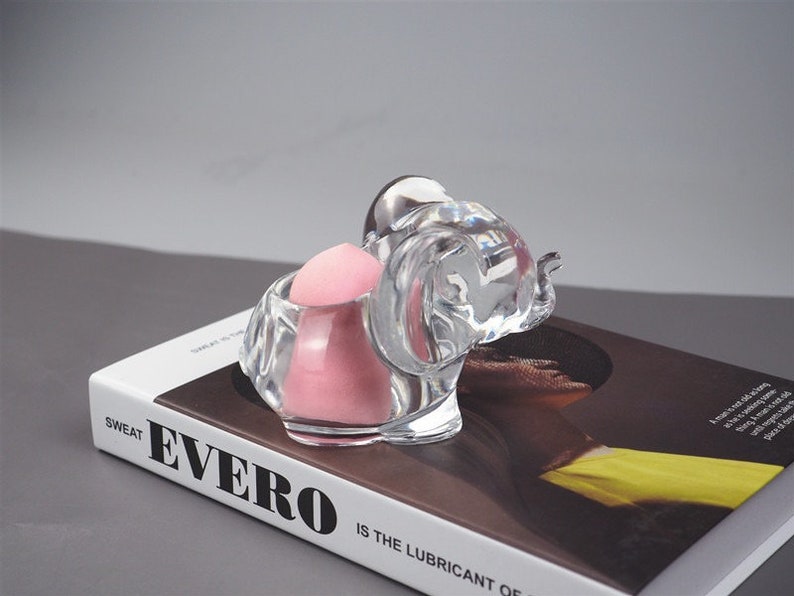 Puede incluir: Una figurita de elefante de cristal transparente con una esponja de maquillaje rosa en su interior. El elefante est&aacute; sentado sobre un libro con la palabra "Evero" impresa en la portada.