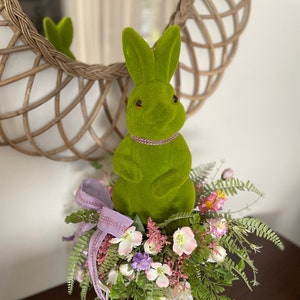 May include: A green bunny ornament with a beaded collar sits on a floral display. The arrangement features white, pink, and purple flowers, green foliage, and a purple ribbon. The bunny is in a white vase with a floral pattern.