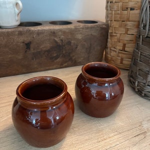 May include: Two small brown ceramic pots with a glossy finish. The pots are sitting on a light wood surface.