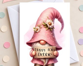 Op de afbeelding: Een wenskaart met een kabouter die een hoge roze muts draagt, versierd met witte madeliefjes. De kabouter houdt een houten bordje vast met de tekst Happy 40th Debbie. De kaart ligt op een witte envelop omringd door gekleurde confetti.