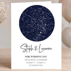 Op de afbeelding: Een witte rechthoekige kaart met een cirkelvormige donkerblauwe sterrenkaart bovenaan. Onder de kaart staat de tekst: Steph & Leeanne, OUR WEDDING DAY, THE RO HOTEL, WINDERMERE, 54.365211 N -2.915630 W, 3RD AUGUST 2024 in zwarte letters.