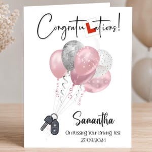 Peut inclure: Une carte de vœux blanche avec le texte Congratulations! incluant un symbole L rouge, un bouquet de ballons roses et argentés, et une clé de voiture. Le nom Samantha est inscrit avec le texte On Passing Your Driving Test et 27/09/2024.