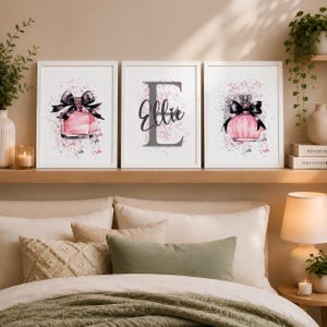May include: A set of three white-framed wall art prints. The left and right prints feature pink perfume bottles with black bows against a pink and grey paint splatter background. The centre print displays a large grey letter E with the name Ellie.
