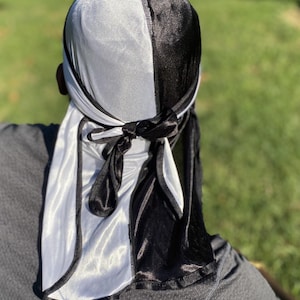 May include: A black and white satin durag with a contrasting colour design. The durag is tied around the head and has a long tail that hangs down the back.