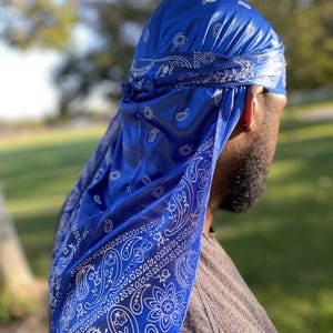 May include: A blue satin bandana with a white paisley pattern. The bandana is tied around a person's head.