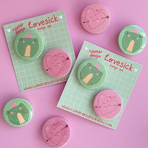 May include: Set of six 1-inch button pins featuring a green frog wearing a crown and a pink heart with an arrow through it. The buttons have the text "Lovesick badge set" and "Buch" on them.