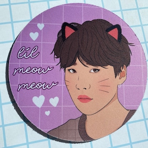 Könnte beinhalten: Ein runder Untersetzer in Lila und Weiß mit einer Cartoon-Illustration einer Person mit braunen Haaren, die Katzenohren trägt. Der Text "lil meow meow" steht auf dem Untersetzer.