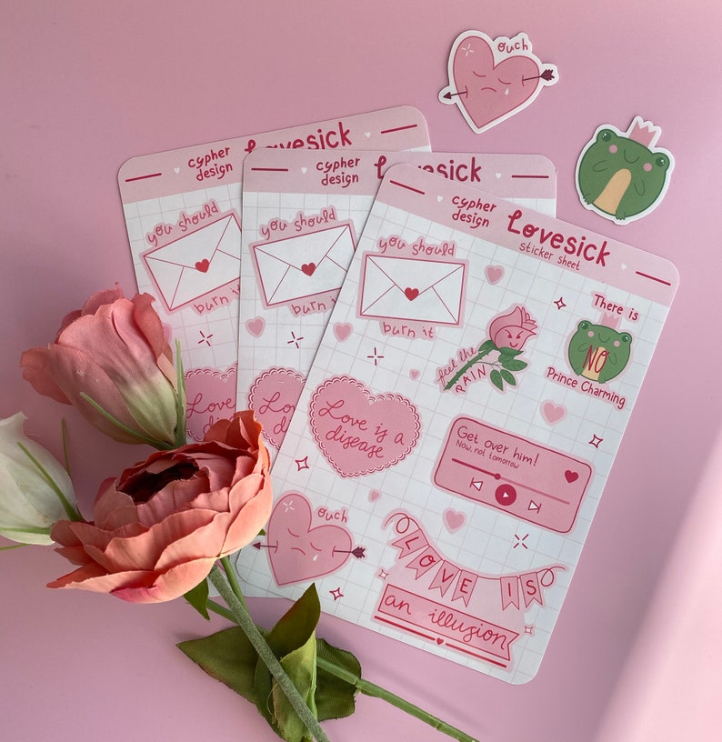 May include: A set of pink and white stickers with various designs, including hearts, flowers, frogs, and text. The text includes phrases like "Love is a disease", "Get over him!" and "Love is an illusion".