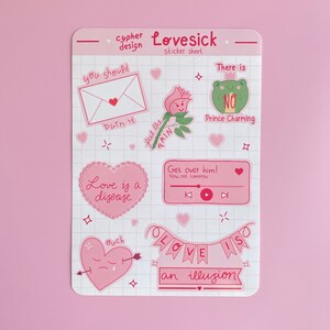 May include: A sheet of pink and white stickers with a love theme. The stickers feature illustrations of a heart with an arrow, a frog, a rose, a music player, and a banner that says "Love is an illusion".