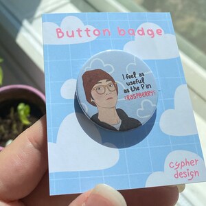 May include: A round button badge with a cartoon illustration of a person wearing a brown beanie and glasses. The text on the badge reads "I feel as useful as the P in "raspberry" ". The badge is on a blue and white background with the text "Button badge" and "cypher design".