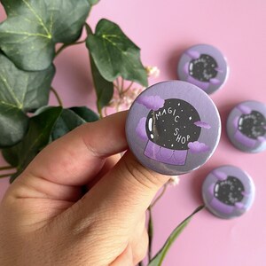 Op de afbeelding: Vier ronde, paarse en zwarte knoppen met een magische winkelthema. De knoppen hebben een glazen bol met sterren en wolken. De tekst op de knop luidt "Magic Shop".
