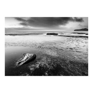 Puede incluir: Una fotografía en blanco y negro de una costa rocosa durante la marea baja. Una gran piedra solitaria descansa en primer plano dentro de un charco poco profundo, mientras las olas rompen contra rocas lejanas bajo un cielo oscuro y nublado.