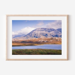 May include: A framed landscape print featuring a mountain range under a blue sky with scattered clouds. The foreground shows a lake and grassy area, with the mountains in the background. The print is in a light wood frame.