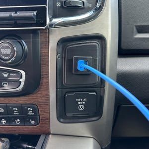 Könnte beinhalten: Nahaufnahme des Armaturenbretts eines Fahrzeugs mit einem USB-Anschluss und einem blauen Kabel. Darunter befindet sich eine 115V AC 150W Steckdose. Das Armaturenbrett hat Holzmaserung und verschiedene Bedienelemente.