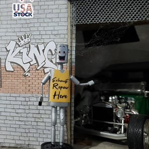 May include: A whimsical metal robot figure with a smiling face and a yellow vest that reads "Exhaust Repair Here." The figure stands in front of a brick wall with graffiti that says "KING." A classic car is visible in the background.