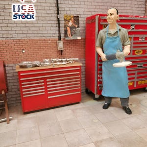 Może przedstawiać: Miniaturowa scena warsztatowa z czerwoną skrzynią na narzędzia i figurką mechanika trzymającego szlifierkę. Figurka ma na sobie niebieski fartuch i szare spodnie. Tło obejmuje ceglaną ścianę i plakat.