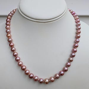 May include: A strand of mauve-colored pearls displayed on a white jewelry bust. The pearls are round and evenly sized, creating a classic necklace design. The necklace is presented against a neutral background.
