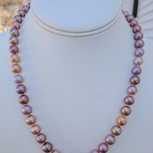 May include: A strand of iridescent, multi-colored pearls displayed on a white bust. The pearls transition in color from pink to bronze and purple. The necklace is a classic jewelry piece.