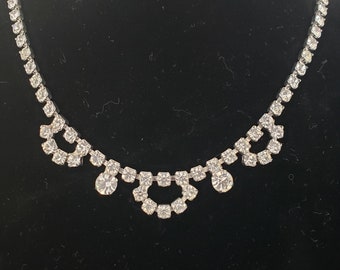 May include: A silver necklace with a delicate design of clear rhinestones. The necklace features a central row of stones with two smaller rows of stones extending out on either side.