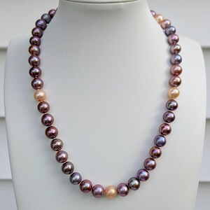 May include: A necklace composed of round, lustrous pearls in varying shades of deep purple, plum, and metallic bronze, interspersed with occasional light peach-colored pearls, arranged in a single strand on a white display bust.