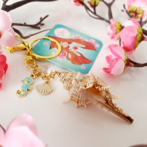 May include: A gold-tone keychain with a seashell charm, a small seashell charm, a seahorse charm, and a pearl charm. The keychain is attached to a key ring.