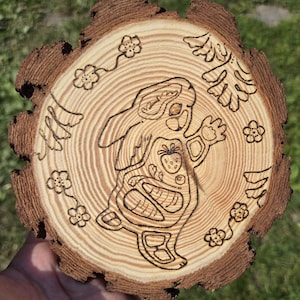 Puede incluir: Una rodaja de madera con un diseño quemado de un conejo de dibujos animados sosteniendo una fresa. El conejo está rodeado de diseños florales y de hojas. La rodaja de madera tiene un borde de corteza natural y rugoso y veta de madera visible.