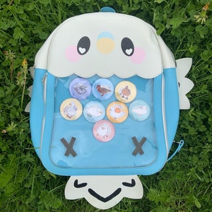 May include: A light blue and white backpack with a bird design, featuring a clear front pocket filled with colorful round pins. The pins have duck illustrations and text. The backpack is on a bed of green grass.