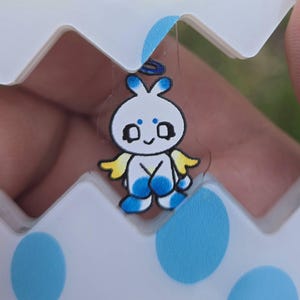 May include: A clear sticker featuring a cartoon character with a white body, blue feet, and yellow wings. The character has a blue halo and is set against a white and blue egg-shaped container with blue polka dots.