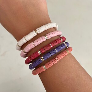 May include: A stack of five colorful beaded bracelets on a person's wrist. The bracelets are made of small, round beads in shades of white, pink, red, purple, and orange. Each bracelet has a thin gold band running through the beads.