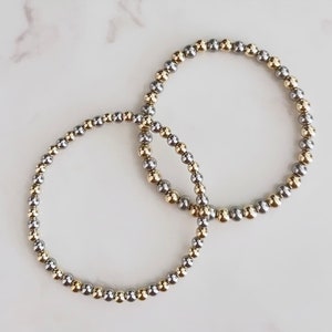 May include: Two bracelets made of silver and gold beads. The bracelets are intertwined and are on a white background.